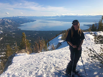 Sierra Nevada University 2021 valedictorian Kayla Heidenrieich stands at the top of a snowy mountain above Lake Tahoe on a sunny day