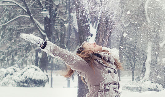 a young woman lifts her face to falling snow among trees